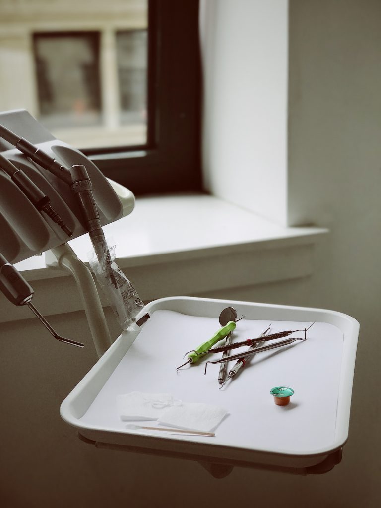 A dental tray with various dental equipment, like dental picks, dental mirrors, suction tubes, and tooth polish sit on a white tray waiting to be used for a dental checkup and cleaning