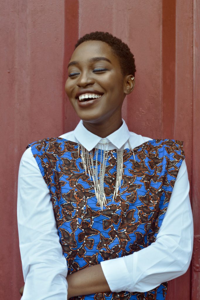 An absolutely stunning, young Black woman has just received a teeth whitening treatment in lafayette, louisiana. she is standing with a red wall behind her and one arm draped across her stomach. She looks happy with a radiant smile.